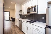 This single-floor unit showcases a clean, contemporary design with a cohesive color palette of white, black, and warm wood tones. Light wood-style flooring runs continuously throughout, complemented by crisp white walls and dark baseboards that create a sharp, modern contrast.

The galley-style kitchen is well-appointed, featuring white shaker cabinetry extending floor to ceiling for ample storage. Stainless steel appliances include a full-size refrigerator, over-the-range microwave, dishwasher, and a black electric cooktop set into white countertops. A dark tile backsplash adds a striking visual accent behind the cooking area, while an open shelf provides additional display or storage space. Recessed lighting illuminates the kitchen corridor, which opens toward a windowed living area at the far end.

The bathroom is finished in a clean, minimalist style with white subway tile surrounding a full soaking tub and shower combination, complemented by light tile flooring and a dark-finished vanity. A separate bedroom area offers a quiet, well-proportioned space equipped with a wall-mounted mini-split HVAC unit for individual climate control. The entry features a wall-mounted shelf and coat hook for convenient organization. Overall, the unit presents a thoughtfully designed, move-in-ready layout.