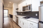 This single-floor unit showcases a clean, contemporary design with a cohesive color palette of white, black, and warm wood tones. Light wood-style flooring runs continuously throughout, complemented by crisp white walls and dark baseboards that create a sharp, modern contrast.

The galley-style kitchen is well-appointed, featuring white shaker cabinetry extending floor to ceiling for ample storage. Stainless steel appliances include a full-size refrigerator, over-the-range microwave, dishwasher, and a black electric cooktop set into white countertops. A dark tile backsplash adds a striking visual accent behind the cooking area, while an open shelf provides additional display or storage space. Recessed lighting illuminates the kitchen corridor, which opens toward a windowed living area at the far end.

The bathroom is finished in a clean, minimalist style with white subway tile surrounding a full soaking tub and shower combination, complemented by light tile flooring and a dark-finished vanity. A separate bedroom area offers a quiet, well-proportioned space equipped with a wall-mounted mini-split HVAC unit for individual climate control. The entry features a wall-mounted shelf and coat hook for convenient organization. Overall, the unit presents a thoughtfully designed, move-in-ready layout.