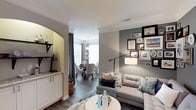 Located at 100 Village Crossing Dr in Chapel Hill, NC, this single-floor residence offers a thoughtfully designed layout that blends comfort and sophistication. The living area features a tufted sectional sofa, a round white coffee table, and a curated gallery wall accented by warm and cool-toned artwork, creating an inviting focal point. Crown molding along the ceiling adds a refined architectural touch throughout the space. A stylish floor lamp with an adjustable arm provides both functional and ambient lighting. An elegant arched entryway transitions seamlessly into the dining area, which is illuminated by a classic chandelier and surrounded by natural light streaming through large windows dressed with floor-length drapes. The dining space features a round wooden table paired with upholstered chairs, ideal for intimate gatherings. Adjacent to the living space, a wet bar or butler's pantry area is visible, showcasing white cabinetry with granite countertops and open shelving — a practical and attractive feature. Dark hardwood flooring runs throughout, grounding the warm neutral color palette of soft grays and whites that defines the overall aesthetic of this well-appointed home.