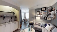 Located at 100 Village Crossing Dr in Chapel Hill, NC, this single-floor residence offers a thoughtfully designed layout that blends comfort and sophistication. The living area features a tufted sectional sofa, a round white coffee table, and a curated gallery wall accented by warm and cool-toned artwork, creating an inviting focal point. Crown molding along the ceiling adds a refined architectural touch throughout the space. A stylish floor lamp with an adjustable arm provides both functional and ambient lighting. An elegant arched entryway transitions seamlessly into the dining area, which is illuminated by a classic chandelier and surrounded by natural light streaming through large windows dressed with floor-length drapes. The dining space features a round wooden table paired with upholstered chairs, ideal for intimate gatherings. Adjacent to the living space, a wet bar or butler's pantry area is visible, showcasing white cabinetry with granite countertops and open shelving — a practical and attractive feature. Dark hardwood flooring runs throughout, grounding the warm neutral color palette of soft grays and whites that defines the overall aesthetic of this well-appointed home.