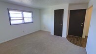 This single-floor residential space offers a straightforward and functional layout designed for comfortable everyday living. The main room features wall-to-wall carpeting in a neutral beige tone, providing a warm and quiet underfoot feel. Light green-tinted white walls keep the space feeling fresh and clean, while white baseboards add a crisp finishing detail throughout. A window fitted with horizontal blinds allows natural light to filter into the room, brightening the interior and offering a view to the outside. Two dark-framed interior doors are prominently positioned along one wall, providing access to additional areas of the space. A transition from carpet to hardwood-style flooring is visible at the edge of the room, suggesting a distinct adjoining area such as a hallway or utility space. The textured ceiling adds a subtle architectural element consistent with traditional residential construction. Overall, the space presents a clean, move-in-ready environment with a neutral palette that offers flexibility for a variety of furnishing and décor styles.
