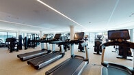 This single-floor fitness center is designed with a contemporary aesthetic, offering a comprehensive and well-organized layout that accommodates a wide range of workout disciplines. The space is divided into several distinct zones, each tailored for specific training needs.

The primary cardio area features an extensive row of Life Fitness treadmills equipped with individual touchscreen monitors, complemented by additional cardio equipment including stair climbers and stationary bikes. Large floor-to-ceiling windows line this zone, allowing natural light to fill the space and providing views of the surrounding cityscape.

A dedicated strength training zone houses a robust selection of selectorized machines, free weights, and cable systems, all finished in sleek black. A mirror wall amplifies the sense of space and supports proper form during training.

A separate functional training room, accessible through full-height glass-panel sliding doors, features a Cascade treadmill, rowing equipment, and open floor space for dynamic exercises, including a heavy-duty tire sled. This area is outfitted with wall-mounted mirrors and a large display screen.

Throughout the facility, speckled terrazzo-style flooring is contrasted by warm wood-tone hardwood accents near entryways. Recessed and linear LED lighting provides consistent, well-distributed illumination across all zones, contributing to a clean, professional atmosphere.