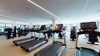 This single-floor fitness center is designed with a contemporary aesthetic, offering a comprehensive and well-organized layout that accommodates a wide range of workout disciplines. The space is divided into several distinct zones, each tailored for specific training needs.

The primary cardio area features an extensive row of Life Fitness treadmills equipped with individual touchscreen monitors, complemented by additional cardio equipment including stair climbers and stationary bikes. Large floor-to-ceiling windows line this zone, allowing natural light to fill the space and providing views of the surrounding cityscape.

A dedicated strength training zone houses a robust selection of selectorized machines, free weights, and cable systems, all finished in sleek black. A mirror wall amplifies the sense of space and supports proper form during training.

A separate functional training room, accessible through full-height glass-panel sliding doors, features a Cascade treadmill, rowing equipment, and open floor space for dynamic exercises, including a heavy-duty tire sled. This area is outfitted with wall-mounted mirrors and a large display screen.

Throughout the facility, speckled terrazzo-style flooring is contrasted by warm wood-tone hardwood accents near entryways. Recessed and linear LED lighting provides consistent, well-distributed illumination across all zones, contributing to a clean, professional atmosphere.