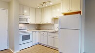 This upgraded 1-bedroom, 1-bathroom unit offers a thoughtfully renovated interior spread across a single floor. The kitchen serves as a standout feature, showcasing white shaker-style cabinetry that extends to the ceiling for generous storage, granite-patterned countertops, and a stainless steel sink. White appliances, including a range with microwave above and a full-size refrigerator, are complemented by warm wood-look luxury vinyl plank flooring and track lighting overhead, creating a bright and functional cooking space. The open layout connects the kitchen to the living area, which transitions to plush carpeting. The living room is generously sized, featuring a sliding glass door that opens to an outdoor balcony with views of surrounding trees, allowing natural light to fill the space. A built-in shelving unit near the entryway provides additional storage. The bedroom is a comfortable, well-proportioned room with soft carpeting and a large window offering pleasant outdoor views. The bathroom features a single-sink vanity with a granite-style countertop, a mirrored medicine cabinet, wood-look tile flooring, and a walk-in shower with built-in shelving and grab bars for added accessibility.
