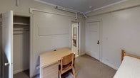 Located at 5540 S Hyde Park Blvd in Chicago's historic Hyde Park neighborhood, this single-floor unit offers a straightforward and functional layout. The bedroom is carpeted and finished with crown molding, featuring a single bed frame, a wooden dresser, and a set of wall-mounted shelves for storage. A window with a roller shade allows natural light into the room while overlooking neighboring brick architecture, and a classic cast-iron radiator provides in-unit heating. A compact secondary area within the room accommodates a mini-refrigerator and microwave, adding convenience for everyday living. A dedicated study nook with a wooden desk and chair provides a practical workspace, with exposed utility piping running along the upper wall lending an utilitarian character to the space. The private bathroom includes a pedestal sink with chrome fixtures, a mirrored medicine cabinet, and a shower enclosed by a white curtain, with tile surround and a skylight overhead that introduces additional natural light. The overall aesthetic is modest and practical, suited for focused, no-frills living in one of Chicago's most storied and intellectually vibrant neighborhoods, in close proximity to the University of Chicago campus.