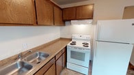 This single-story unit offers a practical and straightforward layout across several well-defined areas. The kitchen is equipped with an electric coil range, a side-by-side configuration with a full-size refrigerator, and an under-cabinet range hood. Warm-toned wood cabinetry lines the upper and lower walls, providing ample storage, while laminate countertops extend along an L-shaped configuration. A stainless steel sink is positioned along the counter, and light vinyl flooring completes the space.

The living area is generously proportioned, featuring wall-to-wall carpet in a neutral tan tone and a sliding glass door fitted with vertical blinds that allows natural light to fill the room. A hallway connects the living space to the rest of the unit, lined with doors leading to additional rooms.

The bedrooms are carpeted and finished with neutral walls, each featuring windows fitted with horizontal blinds that bring in natural light. The rooms offer a clean, open feel with sufficient space for standard furnishings.

The bathroom includes a vanity with a white countertop and integrated sink, a wall-mounted mirror, and decorative tile accenting the wall. Tile flooring and a door opening to the hallway complete the space. Overall, the unit presents a clean, functional living environment.