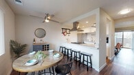 This two-floor residence offers a thoughtfully designed layout with a seamless flow between living spaces. The main level centers around an open-concept kitchen and dining area, where a white peninsula with a built-in cooktop and stainless steel range hood serves as the focal point. The kitchen features white cabinetry, stainless steel appliances, recessed lighting, and a multi-seat breakfast bar with black stools — ideal for casual dining. Adjacent to the kitchen, the dining area accommodates a round wood table and benefits from natural light streaming through a large window fitted with vertical blinds. A ceiling fan overhead adds comfort to the space, while a grey sideboard with a wine rack provides practical storage.

The living room is spacious and open, with rich dark hardwood-style flooring that carries throughout the main level. A white media unit anchors the room, complemented by neutral walls and access to the rest of the home via a central hallway.

Upstairs, the bedroom is a calm retreat featuring plush carpeting, a large window with scenic outdoor views, and striped curtains that filter natural light. The full bathroom includes a tub-shower combination, wood-look flooring, and clean white finishes, while a half bath on the main level offers added convenience.