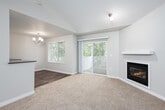 This well-appointed single-floor residence offers a thoughtfully designed open layout that seamlessly connects the living, dining, and kitchen areas. The spacious living room is anchored by a classic fireplace with a clean white surround and plush neutral carpeting underfoot, creating a warm and inviting atmosphere. A sliding glass door opens directly onto a private balcony, framing serene, tree-lined views and allowing generous natural light to fill the space.

The dining area transitions gracefully from the living room, featuring wood-look flooring and an elegant chandelier overhead, with an additional window and balcony door that further enhances the sense of openness and connection to the outdoors.

The kitchen is both functional and stylish, showcasing white shaker cabinetry, a classic subway tile backsplash, and dark countertops that provide a striking contrast. Stainless steel appliances — including a refrigerator, electric range, dishwasher, and mounted microwave — complete the modern aesthetic, while wood-look flooring ties the space together cohesively.

A doll's-eye view of the floor plan reveals a compact, efficiently arranged layout that also includes a bathroom with white cabinetry and additional storage areas. Throughout the residence, a neutral color palette and consistent finishes create a clean, contemporary ambiance.