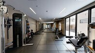 This dedicated weight room in Denver, Colorado offers a well-appointed fitness environment designed for serious strength and conditioning training. The space is laid out efficiently, maximizing every square foot with a thoughtful arrangement of equipment along the perimeter and open floor space in the center. Durable black rubber flooring tiles cover the entire area, providing a safe, impact-resistant surface suitable for heavy lifting and dynamic movement. Linear recessed LED lighting runs across the ceiling, delivering bright, even illumination throughout the room. Large floor-to-ceiling sliding glass windows along one wall flood the space with natural light and provide views to an outdoor area, adding an open, airy quality to the interior. The warm, neutral-toned walls complement the modern aesthetic. Equipment includes a power rack with barbells and weight plates, a full dumbbell rack, cable machines, medicine ball storage, a heavy punching bag, a weight bench, and various selectorized strength machines. A set of double doors with frosted glass panels serves as the entrance, accented by warm wood framing that adds a refined touch. The overall ambiance is clean, functional, and professional, making this weight room a comprehensive facility for a full-body strength training regimen.