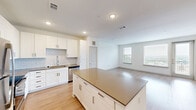 Located at 2001 Plateau Vista Blvd in Round Rock, TX, this one-bedroom, one-bathroom residence offers a thoughtfully designed open-concept layout filled with natural light. The combined kitchen and living area serves as the heart of the home, featuring light hardwood-style flooring that flows seamlessly throughout the space. The kitchen is well-appointed with crisp white shaker cabinetry, a classic subway tile backsplash, stainless steel appliances including a refrigerator, range, microwave, and dishwasher, and a spacious center island with a neutral-toned countertop — ideal for dining or meal preparation. A built-in shelving nook and a dedicated desk area provide additional functional storage and workspace within the living area. Large windows and a glass door open the living space to outdoor views, enhancing the bright, airy ambiance. The bedroom is a comfortable retreat finished with plush carpet, a ceiling fan, and a generous window with outdoor views. The bathroom features light hardwood-style flooring, white cabinetry, neutral countertops, and a glass-enclosed shower, with direct access to a well-organized walk-in closet complete with multi-tier shelving and hanging space. The clean, neutral palette throughout creates a modern and welcoming atmosphere.
