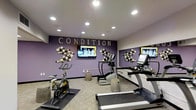 This fitness room is designed with both function and style in mind. The space is anchored by a bold purple accent wall bearing the word "CONDITION" in large lettering, immediately establishing the room's purpose and energetic atmosphere. Decorative circular wall art flanks the signage on either side, adding a creative, modern touch to the interior design.

The room is equipped with a selection of cardio machines, including a stationary spin bike, a treadmill, and an elliptical trainer, providing users with a variety of workout options. Rubber floor mats are positioned beneath the equipment to protect the light-toned flooring and ensure stability during exercise. Cylindrical stool-style seats are placed throughout the space, offering convenient rest points between workouts.

Two wall-mounted flat-screen televisions are positioned for optimal viewing from the equipment, enhancing the workout experience. A large mirror on one wall visually expands the room, reflecting both the equipment and décor. Recessed ceiling lighting provides bright, even illumination throughout the space, contributing to an inviting and motivating environment well-suited for daily fitness routines.