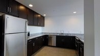 This single-floor space showcases a thoughtfully designed kitchen that serves as its centerpiece. The kitchen features rich, dark espresso-toned cabinetry with sleek hardware, offering generous storage through both upper and lower cabinets arranged in an L-shaped layout. The countertops are finished in speckled granite, providing a striking contrast against the dark cabinetry while offering a durable and elegant workspace. Stainless steel appliances, including a full-size refrigerator, dishwasher, and range, lend a modern, cohesive aesthetic throughout the kitchen. Recessed lighting in the bright white ceiling provides ample, even illumination across the entire space, enhancing the clean and contemporary atmosphere. The neutral-toned flooring complements the overall color palette, tying the design elements together seamlessly. The crisp white walls add to the sense of openness and brightness, creating an inviting environment. The space presents a well-appointed, move-in-ready layout with a clear emphasis on modern finishes and practical design, making it well-suited for everyday living and entertaining alike.