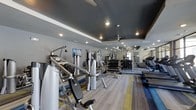 This fitness center is thoughtfully designed to support a complete workout experience. The space features a dark, tray-style ceiling accented with recessed lighting and ceiling fans, creating a modern and energizing atmosphere. The flooring consists of a multi-toned striped carpet that adds a dynamic visual element to the workout environment.

The strength training area is well-stocked with a variety of cable machines, multi-use weight stations, and a full rack of dumbbells organized by weight, catering to users of all fitness levels. Wall-mounted mirrors expand the sense of space and allow for proper form monitoring during exercise.

The cardio section lines the perimeter of the room and includes a row of treadmills and other aerobic equipment, providing ample options for cardiovascular training. Mounted flat-screen televisions are positioned throughout the space to keep users entertained during their workouts.

The layout is open and spacious, allowing for comfortable movement between equipment. The combination of modern finishes, strategic lighting, and a comprehensive equipment selection makes this fitness center a fully functional and visually appealing facility suited for a wide range of exercise routines.