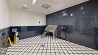 Located at 3010 Edgeview, this 180-square-foot pet grooming room is a purpose-built amenity space designed with both functionality and style in mind. The single-room layout is finished with a striking combination of materials that make it as practical as it is visually appealing. The walls feature glossy navy blue ceramic tile wainscoting that extends approximately halfway up, installed in a classic square pattern on the lower sections and a bold herringbone pattern on the accent wall. Above the tile, the walls transition to a clean, bright white finish, creating a sharp two-tone contrast throughout the space. The floor is covered in a detailed black-and-white checkered mosaic tile, complete with a centrally located floor drain for easy cleanup. The room is equipped with a stainless steel elevated pet wash tub with an integrated step ramp for pet access, along with a mounted grooming hose and spray attachment. A wall-mounted soap or product dispenser and a stainless steel waste bin add to the room's utilitarian design. Recessed ceiling lighting and a ceiling-mounted HVAC unit ensure comfortable conditions year-round. Entry is provided through a glass-panel door with an automatic door closer, offering a clear sightline into the space.