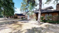 This single-story property presents a classic brick exterior complemented by a dark shingled roof, lending the structure a timeless, traditional aesthetic. The grounds are shaded by several mature, towering pine trees that frame the property and provide natural coverage throughout the yard. The open, expansive yard space surrounding the home offers ample outdoor room, with a concrete pathway leading to the front entrance.

A notable feature of the property is the presence of a separate outbuilding or detached structure positioned to the left of the main building. This additional structure shares the same brick construction and dark roofline, suggesting it may serve as a garage, workshop, or supplementary storage space, adding a layer of functional versatility to the overall property.

Lattice fencing accents are visible along the side of the home, providing subtle decorative detailing and partial privacy screening. The windows on the facade feature light-colored shutters, contributing to the home's traditional character. The overall setting conveys a quiet, established feel, with mature landscaping and a generous lot that together create a private, secluded atmosphere well-suited for comfortable single-story living.