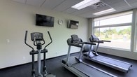 This fitness center offers a clean, functional environment designed to support an effective workout routine. The space is outfitted with multiple Life Fitness cardio machines, including a row of treadmills and an elliptical trainer, providing users with a solid selection of aerobic exercise options. Two wall-mounted flat-screen televisions are positioned for easy viewing from the cardio equipment, allowing users to stay entertained or informed during their sessions. A wall clock is conveniently placed between the screens for time management during workouts. The room features a drop ceiling with integrated fluorescent lighting that ensures consistent, even illumination throughout the space. A large window along one wall allows natural light to fill the room while offering a pleasant view of the surrounding greenery outdoors, contributing to an open and inviting atmosphere. The dark flooring provides a practical, durable surface suited for fitness use, and the light-colored walls keep the space feeling bright and neutral. Overall, this fitness center is thoughtfully arranged to maximize usability while maintaining a clean and comfortable environment for regular exercise.