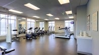 This fitness center offers a well-organized and spacious environment designed to support a variety of cardio-focused workouts. The open floor plan is anchored by durable dark rubber flooring, providing both traction and comfort underfoot. A generous selection of cardio equipment fills the space, including treadmills, elliptical trainers, stationary bikes, and rowing machines, all arranged to allow ample room for movement between stations. Large windows line the walls, flooding the interior with natural light and offering views of the surrounding outdoor environment, creating a bright and energizing atmosphere. Recessed panel lighting in the suspended ceiling supplements the natural light, ensuring consistent illumination throughout all hours of use. A wall-mounted television is positioned for easy viewing during workouts, adding an entertainment element to the exercise experience. Informational and instructional posters are displayed on the walls, offering guidance and motivation to users. Full-length mirrors along one wall enhance the sense of space and allow users to monitor their form. The overall ambiance is clean, functional, and professional, making this fitness center a practical and inviting space for daily exercise routines.