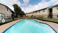This residential community offers a welcoming outdoor environment centered around a spacious swimming pool. The pool features clear, blue water with a classic geometric shape and is finished with a blue tile border, complemented by a surrounding concrete deck ideal for lounging. A sturdy wrought iron fence encloses the pool area, providing both safety and a defined boundary between the recreational space and the well-kept green lawn beyond.

The grounds are generously landscaped, with mature trees offering natural shade and a sense of established character. The multi-unit residential buildings feature a neutral beige exterior with dark trim accents, exterior staircases, and multiple windows that allow ample natural light into each unit. The buildings are arranged to overlook the shared outdoor amenities, creating a cohesive community atmosphere.

The outdoor common area is designed to serve as a central gathering point, balancing functional amenities with open green space. The combination of the pool, manicured lawn, and tree canopy creates a relaxed, resort-like ambiance. The community's layout promotes accessibility to outdoor recreation while maintaining a secure and well-organized environment for its residents.