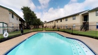 This residential community offers a welcoming outdoor environment centered around a spacious swimming pool. The pool features clear, blue water with a classic geometric shape and is finished with a blue tile border, complemented by a surrounding concrete deck ideal for lounging. A sturdy wrought iron fence encloses the pool area, providing both safety and a defined boundary between the recreational space and the well-kept green lawn beyond.

The grounds are generously landscaped, with mature trees offering natural shade and a sense of established character. The multi-unit residential buildings feature a neutral beige exterior with dark trim accents, exterior staircases, and multiple windows that allow ample natural light into each unit. The buildings are arranged to overlook the shared outdoor amenities, creating a cohesive community atmosphere.

The outdoor common area is designed to serve as a central gathering point, balancing functional amenities with open green space. The combination of the pool, manicured lawn, and tree canopy creates a relaxed, resort-like ambiance. The community's layout promotes accessibility to outdoor recreation while maintaining a secure and well-organized environment for its residents.