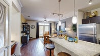 This single-floor residence offers a thoughtfully designed open-concept layout that seamlessly connects the kitchen, dining, and living areas. Rich hardwood-style flooring flows throughout the main living spaces, complemented by crown molding and neutral-toned walls that create a warm, cohesive atmosphere.

The kitchen is a standout feature, equipped with dark espresso cabinetry extending to the ceiling, stainless steel appliances including a refrigerator and microwave, a tile backsplash, and light granite countertops. A peninsula with bar-height seating and stylish pendant lighting bridges the kitchen to the dining area, making it ideal for both everyday use and entertaining.

The open living room benefits from abundant natural light through large windows dressed with dark curtain panels and blinds. A ceiling fan overhead and ample space for comfortable seating add to the room's functionality. Track lighting and recessed fixtures provide additional illumination throughout the main area.

The bedroom is a serene retreat, featuring plush carpeting, a bold accent wall, large windows, crown molding, and a ceiling fan. Generous closet space adds practicality to the room's comfort.

The bathroom offers a clean, modern aesthetic with a dark vanity, dual sinks, a wide mirror, tile flooring, and a bathtub/shower combination, with direct access to the bedroom for added convenience.
