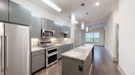 Located at ArtHaus 2828 in Dallas, TX 75204, this one-bedroom, one-bathroom residence offers a thoughtfully designed open-concept layout with contemporary finishes. The combined kitchen and living area serves as the heart of the home, anchored by a chef-inspired kitchen outfitted with sleek gray cabinetry, granite countertops, a striking stone tile backsplash, and a full suite of stainless steel appliances including a gas range, microwave, and side-by-side refrigerator. A generous center island provides additional prep and dining space, while stylish pendant lighting adds a modern touch overhead. Rich wood-tone flooring flows throughout the main living areas, complementing the clean, contemporary aesthetic. The bedroom is a comfortable retreat featuring plush carpeting and a sliding glass door that opens to a private balcony with lush tree views, inviting natural light into the space. A walk-in closet with built-in shelving connects directly to the full bathroom, which includes a soaking tub with tiled surround, granite vanity, and light tile flooring. Additional storage is well-served by a pantry, utility room, and multiple closets throughout.
