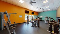 This fitness center makes a strong visual impression with its bold color scheme, featuring striking orange and teal accent walls that energize the space. The single-floor layout is generously proportioned, offering ample room for a comprehensive range of exercise equipment without feeling crowded. Durable rubber flooring covers the entire workout area, providing traction and impact absorption throughout.

The strength training section is well-appointed with multiple selectorized cable machines, a preacher curl station, a leg press, and a flat weight bench, supporting a thorough full-body workout. A dumbbell rack along one wall offers a variety of free weight options. On the cardio side, the space includes several elliptical trainers, treadmills, and a recumbent bike, all positioned for efficient use of the floor plan.

Multiple ceiling fans and a drop-panel ceiling with recessed lighting keep the environment comfortable and well-lit. Wall-mounted flat-screen televisions face the main workout area, adding entertainment during exercise sessions. Large windows along one wall frame views of an outdoor swimming pool and the surrounding grounds, bringing in natural light and an appealing outdoor connection. Mirrors along one section of the wall further enhance the sense of openness while serving a functional purpose for form monitoring.