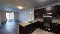 This single-floor space showcases a thoughtfully designed open-concept layout that seamlessly connects the kitchen and living area. The kitchen is a standout feature, equipped with rich, dark espresso cabinetry that provides ample storage and a striking visual contrast against the light granite countertops. A peninsula with a double stainless steel sink extends into the space, offering additional workspace and a natural division between the kitchen and living areas. Pendant lighting hangs above the peninsula, adding a modern, decorative touch. The kitchen is fully appointed with black appliances, including a range and dishwasher, as well as a stainless steel microwave, and is finished with a stylish tile backsplash. Wood-look flooring runs throughout, lending warmth and continuity to the space. The open living area beyond the kitchen benefits from natural light streaming in through a large sliding glass door, while a ceiling fan adds comfort and functionality. The clean white walls and modern lighting fixtures contribute to a bright, contemporary ambiance, making this a practical and visually cohesive space well-suited for everyday living.