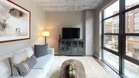 This single-floor unit showcases a distinctive industrial-modern aesthetic throughout, defined by exposed concrete ceilings and structural columns that contrast with warm, neutral-toned walls and wood-look flooring. The open-concept layout connects the living, dining, and kitchen areas into a cohesive, functional space.

The living area is anchored by large floor-to-ceiling windows with black metal frames, flooding the room with natural light and offering views of the surrounding urban streetscape. A plush area rug, comfortable seating, and a media console contribute to the room's relaxed yet stylish atmosphere.

Adjacent to the living space, the dining area accommodates a four-seat table and is accented by a decorative circular mirror wall display and lush greenery, adding visual interest to the corner.

The kitchen features white cabinetry with matte black hardware, dark countertops, a classic subway tile backsplash, and stainless steel appliances, including a range, microwave, and dishwasher — combining practicality with a clean, contemporary look.

Two bedrooms offer restful retreats, each with exposed concrete ceilings, neutral tones, and warm ambient lighting. One bedroom features a leather headboard and dark hardwood flooring, while the other includes an ornate metal-frame bed with soft white linens.

The bathroom is finished in light tones with a wide vanity, backlit mirror, and a tub-shower combination with a curtain rod and niche shelving.