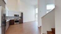 This contemporary residence showcases a thoughtfully designed open-concept layout that makes excellent use of its single floor. Soaring double-height ceilings in the living area create an impressive sense of volume and airiness, complemented by large floor-to-ceiling windows and a glass door that flood the space with abundant natural light and offer sweeping views of the surrounding neighborhood, including glimpses of palm trees in the distance.

The kitchen is a standout feature, outfitted with a full suite of stainless steel appliances including a refrigerator, gas range, built-in microwave, and dishwasher. Two-tone cabinetry — warm wood-toned lower cabinets paired with sleek white upper cabinets — adds visual depth, while a chevron-patterned tile backsplash and light stone countertops deliver a refined, modern aesthetic. Warm-toned hardwood flooring flows seamlessly throughout the main living areas, tying the open spaces together cohesively.

The bathroom features a rich wood vanity with stone countertop and a backlit LED mirror, combining functionality with a spa-like ambiance. Practical conveniences include an in-unit stacked washer and dryer tucked neatly behind louvered doors. Crisp white walls throughout the residence enhance the bright, clean atmosphere, making this a polished and move-in ready space.