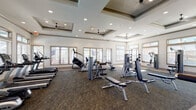 This single-story fitness center is thoughtfully designed to accommodate a wide range of workout needs. The main exercise area is anchored by an impressive coffered ceiling with multiple ceiling fans and recessed lighting, lending the space an open, airy feel. Large windows line the perimeter walls, allowing abundant natural light to fill the room while offering views of the surrounding exterior. The patterned carpet flooring throughout this zone supports an array of strength and cardio equipment, including cable machines, weight benches, selectorized machines, a StairMaster, treadmills, and a dumbbell rack positioned along a floor-to-ceiling mirror. A separate studio room, finished with light hardwood-style flooring and its own coffered ceiling, provides a bright and versatile environment for group fitness activities. This room is outfitted with spin bikes, stability balls, resistance bands, and a wall-mounted television, with glass doors that open to the outdoors. The overall ambiance across both spaces is clean, modern, and well-lit, creating an inviting environment for varied fitness pursuits.