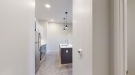 Located at 1625 S 14th St in St. Louis, Missouri, this single-floor unit offers a thoughtfully designed layout with modern finishes throughout. The kitchen is a standout feature, equipped with stainless steel appliances including a refrigerator, range, and microwave, complemented by two-tone cabinetry in white and dark gray. A central island with a white quartz countertop and undermount sink serves as both a prep area and a focal point, illuminated by stylish pendant lights overhead. A textured tile backsplash adds visual interest behind the range. Polished concrete floors extend through the main living areas, lending an industrial-modern character to the space. The open living area benefits from oversized windows dressed with floor-length dark curtains, allowing for adjustable natural light. A stepped transition leads into the bedroom, which features carpeted flooring and a ceiling fan. The bathroom is clean and contemporary, with a white vanity, undermount sink, large mirror, and a glass-enclosed shower with tile surround. A hallway connects the living spaces efficiently, with a bold black entry door providing a striking contrast to the bright white walls throughout the unit.
