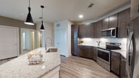 This single-floor residence showcases a thoughtfully designed open layout with consistent, contemporary finishes throughout. The kitchen serves as the centerpiece of the living space, featuring rich dark-espresso cabinetry that extends to the ceiling, a classic white subway tile backsplash, and granite countertops. A generously sized island with an undermount sink and a sleek pendant lighting duo overhead provides both additional workspace and an informal gathering area. Stainless steel appliances, including a refrigerator, range, microwave, and dishwasher, complete the well-appointed kitchen. Wood-look plank flooring runs throughout the main living areas, lending warmth and visual continuity. The living area is bright and open, benefiting from natural light through glass patio doors and a ceiling fan overhead. Bedrooms are carpeted for comfort and include windows that allow natural light. Two bathrooms mirror the kitchen's design language, each with dark cabinetry, granite countertops, large framed mirrors, and wood-look flooring. The primary bathroom includes both a soaking tub and a separate glass-enclosed shower with tile surround. A secondary bathroom offers a tub-shower combination with similar finishes, maintaining a cohesive aesthetic throughout the entire residence.