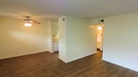 This single-floor residence offers a clean, functional layout designed for comfortable everyday living. The main living area features rich, dark hardwood-style flooring that extends throughout, providing a warm and cohesive aesthetic. A ceiling fan with a light fixture is installed overhead, ensuring air circulation and ambient lighting in the living space. The room is defined by crisp white walls and clean baseboards, contributing to a bright and neutral atmosphere. A partial wall divider separates the living area from the kitchen, creating a sense of distinction between spaces while maintaining an open feel. The kitchen is equipped with white cabinetry, a dishwasher, and countertops with warm-toned finishes, visible through the open pass-through design. Overhead ventilation is present throughout, with ceiling vents ensuring consistent airflow. Adjacent to the main living area, a hallway leads to additional rooms bathed in warm light, suggesting well-connected private spaces. The overall ambiance of the residence is clean, move-in ready, and practical, with a neutral palette that provides a versatile canvas for personalization.