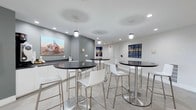 This contemporary common area is thoughtfully designed to serve as a welcoming social hub. The space features a clean, modern aesthetic defined by crisp white walls accented with soft gray tones, creating a bright and open atmosphere. Large-format light gray floor tiles span the entire area, adding a sleek, cohesive foundation to the room.

Several round high-top tables with polished dark granite surfaces are paired with modern white and chrome bar-height chairs, offering comfortable seating arrangements ideal for casual conversation or informal gatherings. Pendant lighting fixtures hang above the space alongside recessed ceiling lights, providing warm, layered illumination throughout.

A built-in counter with dark granite surfaces lines one side of the room, complementing the design of the high-top tables and providing additional surface space. The walls are adorned with a curated selection of large-format coastal and nature-inspired photographic artwork, lending the space a vibrant and scenic character.

A white cabinetry unit is integrated along the wall, adding both functional storage and a clean visual element. The overall design is minimalist yet inviting, balancing functionality with an aesthetic that encourages residents or visitors to linger and connect.