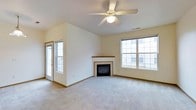 This single-floor space centers around a spacious living area designed for both comfort and functionality. The room features vaulted ceilings that enhance the sense of openness, complemented by a ceiling fan with a built-in light fixture that provides both air circulation and ambient illumination. Soft, neutral-toned carpeting covers the floor throughout, contributing to a warm and inviting atmosphere.

A standout feature of the living area is the fireplace, framed by a classic tile surround and a simple mantel, serving as a natural focal point for the room. Multiple windows, including a large multi-pane window and an additional window near the entry door, allow generous natural light to fill the space and provide views of the surrounding exterior. The entry door with its own window panel adds to the brightness while offering convenient access.

The walls are finished in a clean, neutral cream tone that pairs well with the wood-accented baseboards, creating a cohesive and understated aesthetic throughout. The overall layout feels open and versatile, well-suited for a variety of furniture arrangements and uses.