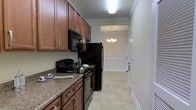 This single-floor residence offers a thoughtfully designed layout that blends functionality with classic style. The kitchen is a standout feature, showcasing rich, warm-toned wood cabinetry with brushed hardware and granite countertops that add a touch of elegance. Black appliances, including a range, over-the-range microwave, and refrigerator, provide a bold contrast against the lighter surroundings. The kitchen flows seamlessly toward the adjoining dining area, which is illuminated by a decorative chandelier that adds a refined, welcoming ambiance. Crown molding accents the transition between spaces, lending an air of architectural detail throughout. Light-toned tile flooring runs through the main areas, contributing to a clean and open feel. The walls are finished in soft, neutral tones that enhance the brightness of the space. Flush ceiling lighting in the kitchen ensures ample illumination for meal preparation, while the overall layout is efficient and well-organized. The interior entrance, visible from the kitchen, features a white paneled door and wainscoting details, further reinforcing the classic design sensibility that carries throughout this well-maintained single-level home.