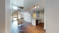 Located at 4605 Cahuenga Blvd in Los Angeles, CA, this two-story space offers a well-designed layout with distinctive architectural character. The main level features rich hardwood flooring throughout, anchored by a striking spiral staircase with carpeted treads and a white metal railing that serves as a visual centerpiece. The living area boasts soaring double-height ceilings that create an impressive sense of openness, complemented by a classic white-framed fireplace and vertical blinds that allow generous natural light to filter in. The kitchen is outfitted with gray shaker-style cabinetry, light quartz countertops, a stainless steel sink, and a full suite of stainless steel appliances including a gas range, refrigerator, and dishwasher, all illuminated by track lighting overhead. The bathroom on the main level includes a tiled shower-tub combination with chrome fixtures and wood-look flooring. Upstairs, the second floor opens to a bright landing with a clerestory window offering neighborhood views. The upper-level rooms feature neutral carpet, crisp white walls, and vertical blinds, providing versatile and comfortable living spaces throughout.