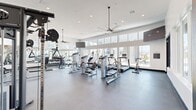This modern fitness center is thoughtfully designed across a single floor, offering two distinct workout zones that together provide a comprehensive exercise environment. The main gym area features soaring ceilings with large clerestory windows that flood the space with natural light, creating an open and energizing atmosphere. Ceiling fans are strategically placed throughout to maintain airflow and comfort during workouts. The durable dark rubberized flooring supports a wide array of strength and cardio equipment, including treadmills, ellipticals, stationary bikes, cable machines, a Smith machine, weight benches, and a full dumbbell rack. Bold motivational murals — including "Hustle for That Muscle" and "Inhale Peace, Exhale Stress" — adorn the walls, adding character and inspiration to the space. Large windows along the perimeter offer views of the surrounding exterior, reinforcing the bright and welcoming environment. The second workout zone features light wood-style flooring, floor-to-ceiling mirrors, a mounted television, and a rowing machine, making it well-suited for yoga, stretching, or group fitness activities. Together, the two areas offer a well-rounded facility designed to support a full spectrum of fitness goals.