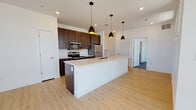 This contemporary single-floor residence showcases a clean, modern design throughout. The open-concept kitchen is a standout feature, centered around a large white quartz island with an undermount sink and seating. Rich dark wood cabinetry lines the kitchen walls, complemented by stainless steel appliances including a refrigerator, electric range, and microwave. Three sleek black pendant lights with gold interiors hang above the island, adding a refined touch. Light wood-tone flooring flows seamlessly from the kitchen into the open living area, which is bathed in natural light from large windows fitted with horizontal blinds. A ceiling fan overhead adds both function and a modern aesthetic to the living space.

The bedrooms feature plush grey carpeting and generously sized windows that offer expansive outdoor views, filling each room with natural light. One bedroom connects directly to a well-appointed bathroom featuring a dual-sink vanity with blue-grey cabinetry, a lighted mirror, light wood flooring, and a full soaking tub with shower. A second bathroom provides a single-sink vanity with wood-toned cabinetry, a lighted mirror, and light wood flooring. Throughout the unit, crisp white walls and recessed lighting contribute to a bright, airy ambiance that feels both modern and welcoming.