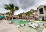 Located at 1215 E Owassa Rd in Edinburg, Texas, this outdoor amenity area offers a resort-style pool experience designed for relaxation and recreation. The centerpiece is a generously sized swimming pool with a shallow wading edge, complemented by an adjacent spa with bubbling water features. Tall palm trees are strategically placed throughout the pool deck, providing a tropical aesthetic and natural shade. The surrounding concrete deck is well-appointed with lounge chairs, colorful seating, and vibrant umbrellas, creating a welcoming atmosphere for outdoor gatherings. Covered pergola structures with stone columns line portions of the deck, offering shaded retreats from the South Texas sun. Wrought iron fencing defines the perimeter of the pool area, providing security while maintaining an open, airy feel. Lush landscaping borders the space with dense shrubs, ornamental palms, and mature trees, enhancing privacy and greenery. The exterior architecture of the surrounding multi-story buildings features a combination of stone and wood-tone siding with covered balconies, contributing to a cohesive residential community aesthetic. A dedicated covered pavilion area with tables and seating provides an ideal space for outdoor dining and socializing, rounding out this well-designed communal outdoor environment.