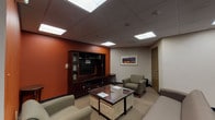 This amenity floor offers a comprehensive range of shared facilities thoughtfully arranged across a single level, accessible via elevators and well-lit corridors with wood-look flooring. The common lounge serves as a central gathering space, featuring warm terracotta-orange accent walls, carpeted floors, comfortable upholstered seating, a dark wood entertainment center with a flat-screen TV, and framed artwork — creating an inviting, relaxed atmosphere. The fitness center is outfitted with a multi-station strength machine, stationary bike, treadmills, and exercise mats, set against sage-green walls under recessed panel lighting. The business center provides desktop computers, a printer, and a small seating area with a round table, making it suitable for work and study. The laundry facility is fully equipped with a long row of front-loading washers and dryers, stacked dryer units, a payment kiosk, and tile flooring for easy maintenance. A communal kitchen area features wood cabinetry, a microwave, dishwasher, and sink for resident use. Together, these amenities are finished consistently with drop ceilings, panel lighting, and neutral tones, delivering a functional and cohesive environment.