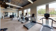 Located on Ellinwood Street in Des Plaines, Illinois, this single-floor fitness center offers a comprehensive workout facility designed to accommodate a wide range of exercise needs. The space features an open, industrial-style aesthetic, with an exposed ceiling showcasing ductwork and mechanical systems alongside sleek linear pendant lighting that illuminates the facility throughout.

The cardio zone is lined with large floor-to-ceiling windows that flood the area with natural light and provide views of the surrounding streetscape, creating an energizing environment. This section houses a row of treadmills, elliptical trainers, and stationary bikes arranged for easy access and comfortable spacing between equipment.

The strength training area is equipped with a power rack, adjustable bench press, dumbbell rack, and a multi-functional cable machine, catering to users at various fitness levels. A dedicated functional training zone features a patterned accent wall, wood-toned flooring, a wall-mounted television, and a mirrored wall section, making it suitable for stretching, cycling, or group-style workouts. Additional storage cubbies and organizational fixtures are integrated throughout the space, maintaining a clean and functional layout. The durable, tile-style flooring runs consistently across the facility, tying the zones together cohesively.