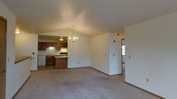 This single-floor space is defined by its open, airy layout and vaulted ceilings that add a sense of generous vertical space to the main living area. A decorative chandelier serves as a focal point, casting warm light across the room and complementing the neutral-toned carpeting that extends throughout the living space.

The kitchen is seamlessly integrated into the open floor plan, separated by a partial peninsula that doubles as a breakfast bar. Warm wood cabinetry lines the kitchen walls, paired with under-cabinet lighting that illuminates the countertops below. A white appliance suite, including what appears to be a range and dishwasher, provides a clean contrast to the rich cabinetry tones. A sink is positioned along the counter, and recessed lighting above ensures the kitchen remains well-lit.

A hallway visible from the living area suggests additional rooms beyond, with natural light filtering in from that direction. The walls throughout are finished in a clean, neutral white, contributing to the bright and open ambiance of the space. Overall, the layout promotes a natural flow between the kitchen and living areas, making it a functional and cohesive environment.