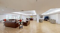 Located at 100 Cole Ln in Lawrence Township, NJ, this single-floor community space offers a generously sized, open-plan layout designed for social gathering and recreation. The main area features light hardwood flooring throughout and elegant tray ceilings with recessed lighting, lending the space a bright, welcoming ambiance. Multiple seating arrangements with plush leather sofas and accent chairs define distinct lounge zones, while large windows along the perimeter fill the interior with natural light and views of the surrounding grounds. A dedicated recreation corner houses a full-size billiards table alongside bench seating, making it an ideal spot for leisure activities. A separate dining area accommodates a round table with chairs and is complemented by a digital piano, adding a refined, communal touch. Interior pass-through windows connect this dining nook to the adjacent galley-style kitchen, which features warm wood cabinetry, dark granite countertops, a double sink, and ample storage — well-suited for catering and event preparation. A media area with a flat-screen television and fireplace provides a cozy retreat, with glass doors opening to an outdoor space. The overall design balances functionality with comfort, making it a versatile venue for a wide range of community activities.