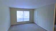 This single-floor residential space offers a clean, functional layout with a consistent design aesthetic throughout. The interior is finished with wall-to-wall carpeting in a neutral tone, providing a comfortable and quiet underfoot feel that complements the soft, warm beige walls. Large windows with horizontal blinds allow ample natural light to fill the space, while offering views of mature trees outside, creating a pleasant connection to the outdoors. The ceilings are smooth and painted white, contributing to a bright and open atmosphere. A standard interior door provides access to additional areas of the unit, and the straightforward, uncluttered layout allows for flexible furniture arrangement. Electrical outlets are conveniently positioned throughout, and a ceiling vent indicates climate control support. The overall ambiance is calm and understated, well-suited for comfortable everyday living. The neutral palette and practical finishes make it an easy canvas for personalization, while the natural light and outdoor greenery visible through the windows add a welcoming warmth to the interior environment.