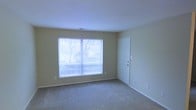 This single-floor residential space offers a clean, functional layout with a consistent design aesthetic throughout. The interior is finished with wall-to-wall carpeting in a neutral tone, providing a comfortable and quiet underfoot feel that complements the soft, warm beige walls. Large windows with horizontal blinds allow ample natural light to fill the space, while offering views of mature trees outside, creating a pleasant connection to the outdoors. The ceilings are smooth and painted white, contributing to a bright and open atmosphere. A standard interior door provides access to additional areas of the unit, and the straightforward, uncluttered layout allows for flexible furniture arrangement. Electrical outlets are conveniently positioned throughout, and a ceiling vent indicates climate control support. The overall ambiance is calm and understated, well-suited for comfortable everyday living. The neutral palette and practical finishes make it an easy canvas for personalization, while the natural light and outdoor greenery visible through the windows add a welcoming warmth to the interior environment.
