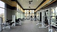 This single-floor fitness center delivers a dynamic and energizing workout environment designed to accommodate a variety of exercise needs. The space is defined by its impressively high ceilings, which are complemented by large floor-to-ceiling windows along the perimeter wall, flooding the interior with natural light and offering views of the surrounding exterior. A bold, striking color scheme of black, gray, and gold adorns the upper walls in a geometric stripe pattern, lending the space a modern and motivating aesthetic. Ceiling fans are mounted overhead to ensure comfortable air circulation throughout. The flooring consists of durable rubber gym material, ideal for both cardio and strength training activities. A row of cardio machines, including treadmills and ellipticals, is centrally positioned, while strength and resistance equipment lines the sides of the room. Large wall-mounted mirrors along one side visually expand the space and allow users to monitor their form during workouts. Exposed ductwork along the upper walls adds an industrial touch that complements the contemporary design. The overall ambiance is clean, well-organized, and professional, making it a functional and inviting facility for fitness enthusiasts.