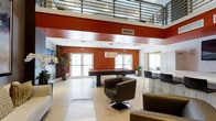 This contemporary community lounge is designed to impress with its dramatic double-height ceilings and an open, airy layout that immediately commands attention. A bold terracotta-red accent wall serves as the visual centerpiece of the space, creating a warm yet energetic backdrop that contrasts beautifully with the crisp white walls and light-toned tile flooring throughout. An upper-level mezzanine with sleek metal railings overlooks the main floor, adding architectural depth and a sense of grandeur to the interior.

The lounge is thoughtfully appointed for both recreation and relaxation. A full-size billiards table anchors the central area beneath pendant lighting, making it an ideal focal point for social gatherings. Nearby, a high-top bar counter with modern bar stools provides a casual space for conversation or work. Comfortable swivel lounge chairs in rich dark leather are arranged throughout, inviting residents to unwind in style.

Natural light floods the space through multiple large glass doors and windows, enhancing the open, welcoming feel. Decorative potted plants and floral arrangements add organic warmth to the modern aesthetic. The combination of recreational amenities, sophisticated design elements, and abundant natural light makes this lounge a well-rounded and inviting communal destination.