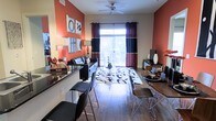 This single-floor residence showcases a vibrant, open-concept layout that seamlessly integrates the dining and living areas into one cohesive space. The interior is defined by bold terracotta-red accent walls contrasted against neutral tones, creating a warm and energetic atmosphere throughout.

The living area centers around a black leather sofa positioned atop a graphic black-and-white area rug, anchoring the space with a modern aesthetic. A ceiling fan overhead provides both functionality and a clean architectural detail. Wall art is thoughtfully arranged on the accent wall, adding a curated, gallery-like feel to the room.

Adjacent to the living area, the dining space features a sleek wooden table paired with contemporary chairs, ideal for everyday meals or entertaining. A bar-height counter with matching stools provides an additional seating option, enhancing the versatility of the layout.

Hardwood-style flooring extends throughout, adding continuity and warmth underfoot. A large sliding glass door floods the interior with natural light and provides direct access to an outdoor area, offering views of surrounding greenery. Dark curtain panels frame the door, adding a dramatic finishing touch to the overall design.