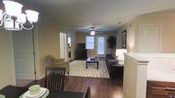 This single-floor residence offers a thoughtfully designed open-concept layout that seamlessly connects the dining and living areas, creating a welcoming and functional environment. Rich, dark hardwood flooring runs throughout, adding warmth and elegance to the space. The living room is a central highlight, featuring a ceiling fan with a light fixture, classic crown molding, and neutral-toned walls that lend a clean, timeless aesthetic. A large window fitted with blinds allows generous natural light to fill the room, enhancing its bright and airy feel. A glass-paneled exterior door provides additional light and access to the outdoors. The living area is spacious enough to accommodate a full seating arrangement, complete with a sofa, accent chair, and a coffee table atop a patterned area rug that anchors the space. A media console with a flat-screen television is positioned along one wall, adding to the room's practicality. An adjoining dining area with dark wood furniture is visible in the foreground, offering a defined yet open space for meals. Framed wall art and decorative accents throughout contribute to a polished, lived-in atmosphere. A hallway visible from the main area suggests additional rooms beyond, rounding out this comfortable and cohesive single-level home.